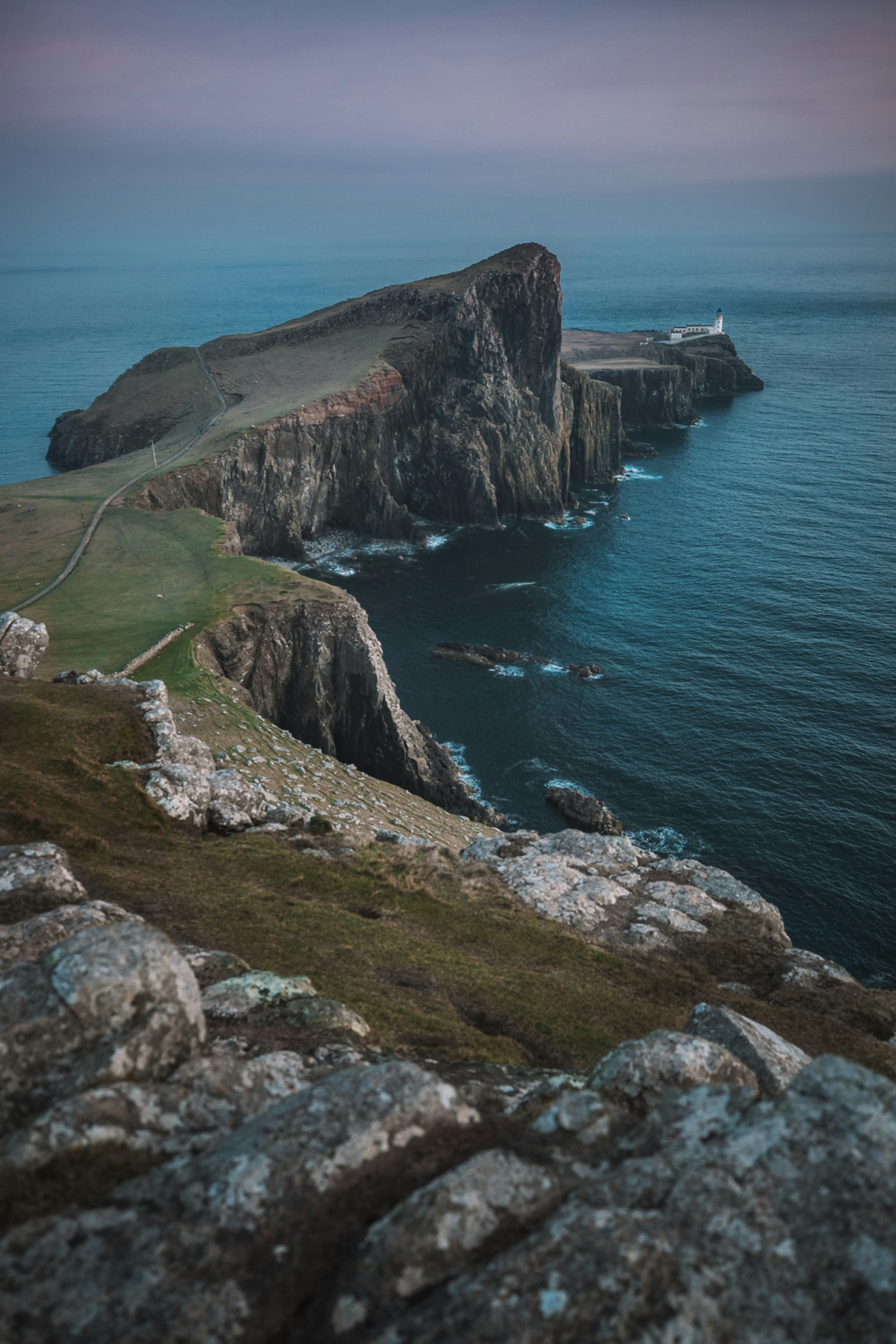 Looking out to Neist Point Lighthouse on the Isle of Skye, Scotland in the evening; it sits at the end of a long promontory of high rocky cliffs topped by grass.
