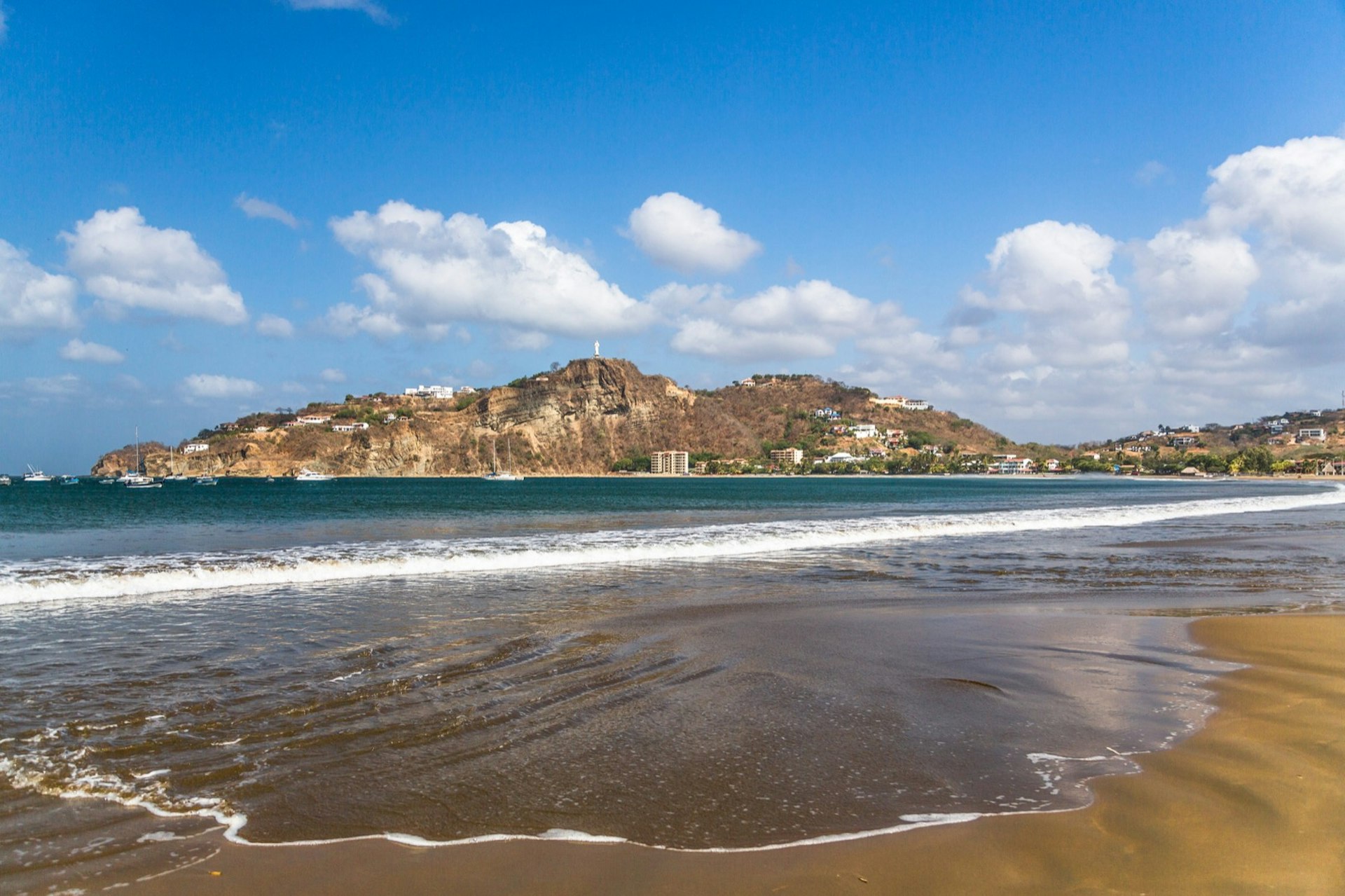 Gentle waves lap the sandy light brown shores. In the background there's a large mountain dotted with homes and hotels. In the distance, a few boats are moored in the ocean; Nicaragua travel. 