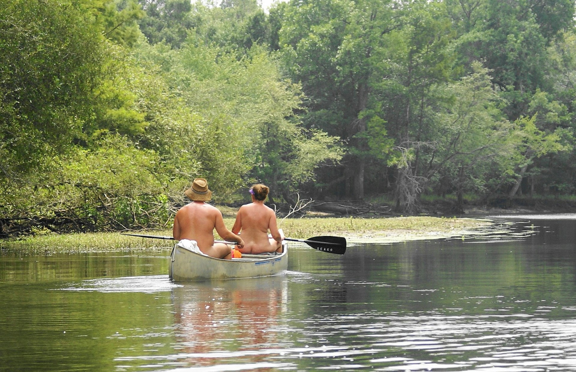 A nude man and woman ride in a canoe down a body of water. There are trees having over the water; nude vacations 