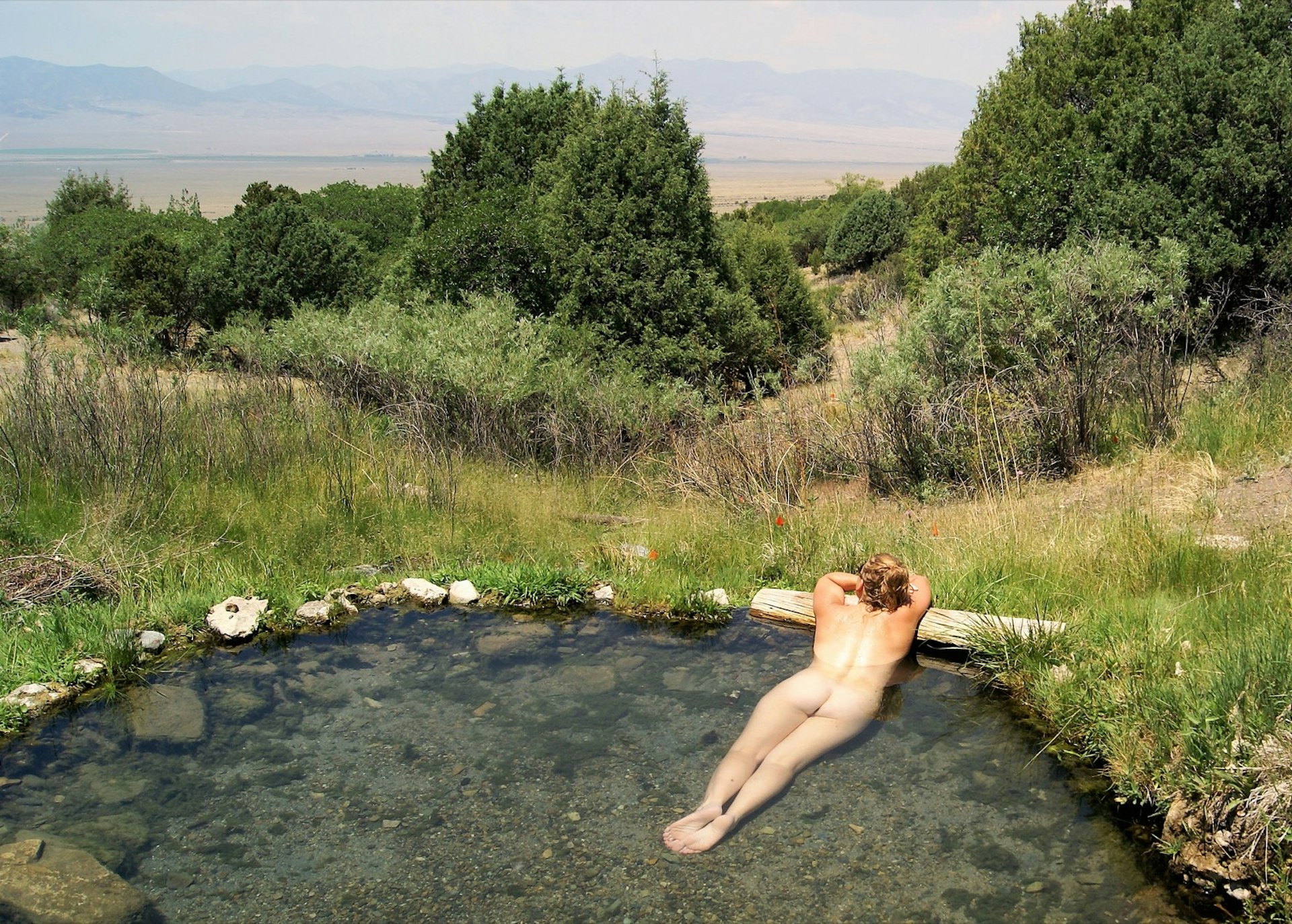 A nude woman relaxes in a small pool of water. She's looking out at green trees and tall bushes; nude vacations 