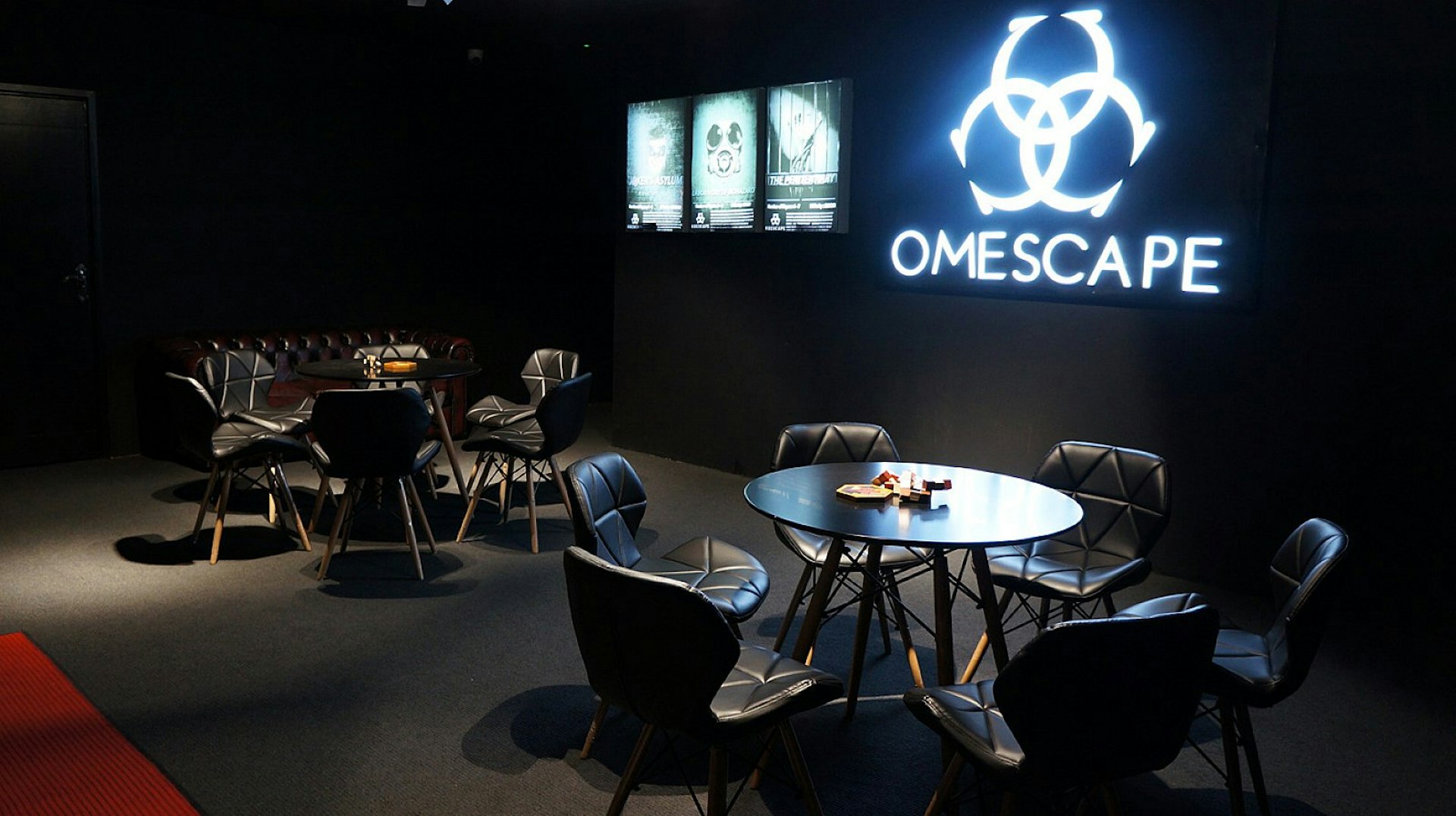 The interior of a dark room, with leather chairs arranged around tables; on the wall is a sign saying Omescape, below a stylised biohazard symbol.