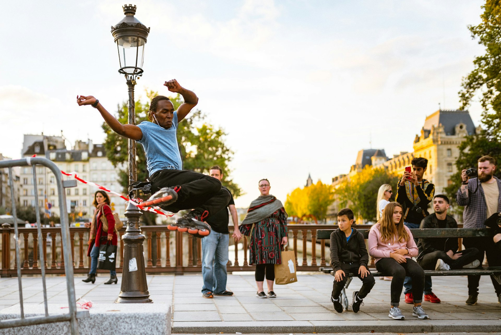A rollerblader jumps some tape on a bridge over a river in Paris; there is an intricate lamppost behind him, and crowds milling around a bench watching; in the background are trees and a handsome building.