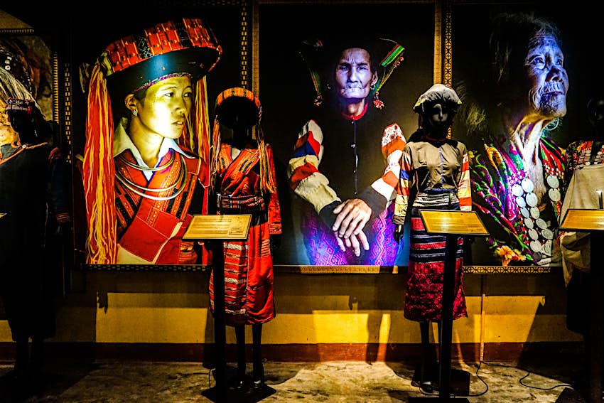 Two mannequins wearing traditional clothing stand in front of three large portraits of women in similar traditional clothing