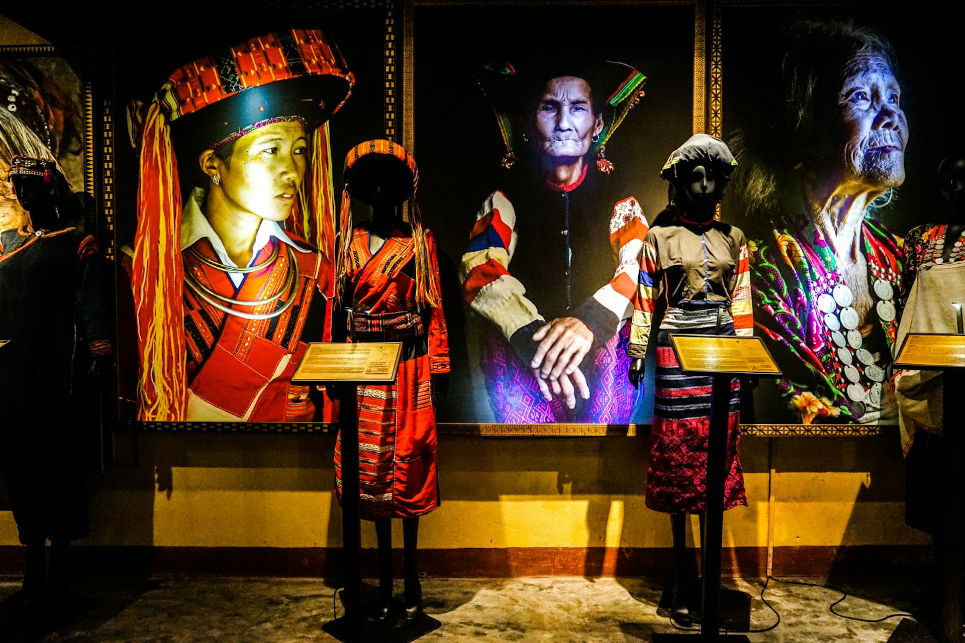 Two mannequins wearing traditional clothing stand in front of three large portraits of women in similar traditional clothing