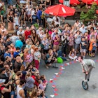 An acrobat stands on a unicycle in a funny pose as a crowd of spectators surround him, sitting on the ground