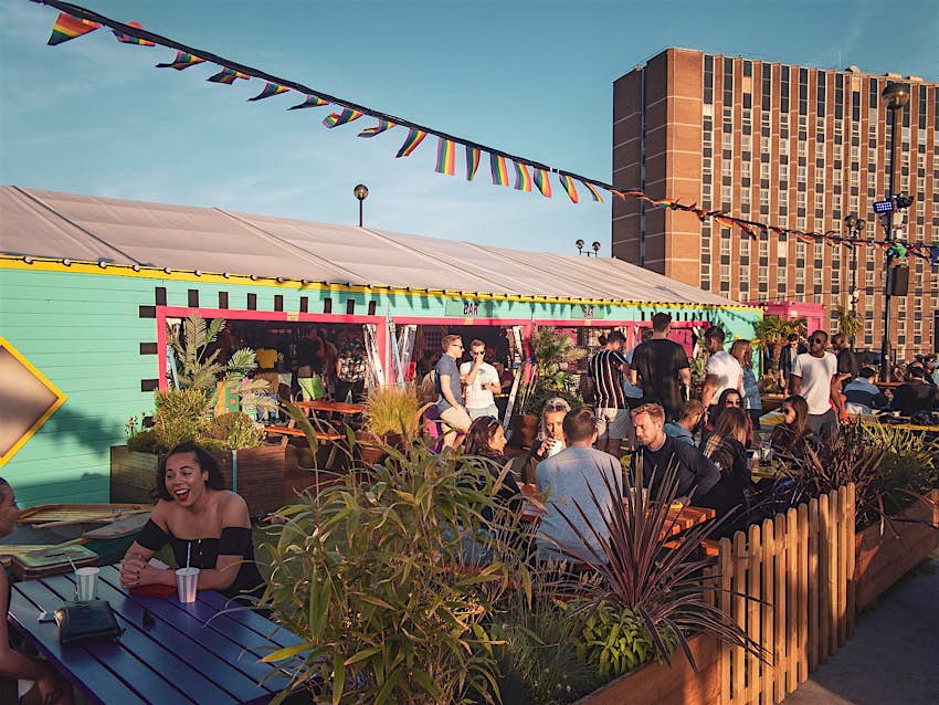 Drinking at wooden benches in the sun on Roof East, under rainbow-coloured bunting; the bar is housed in a bright turquoise structure, with a red-brick office building visible beyond.