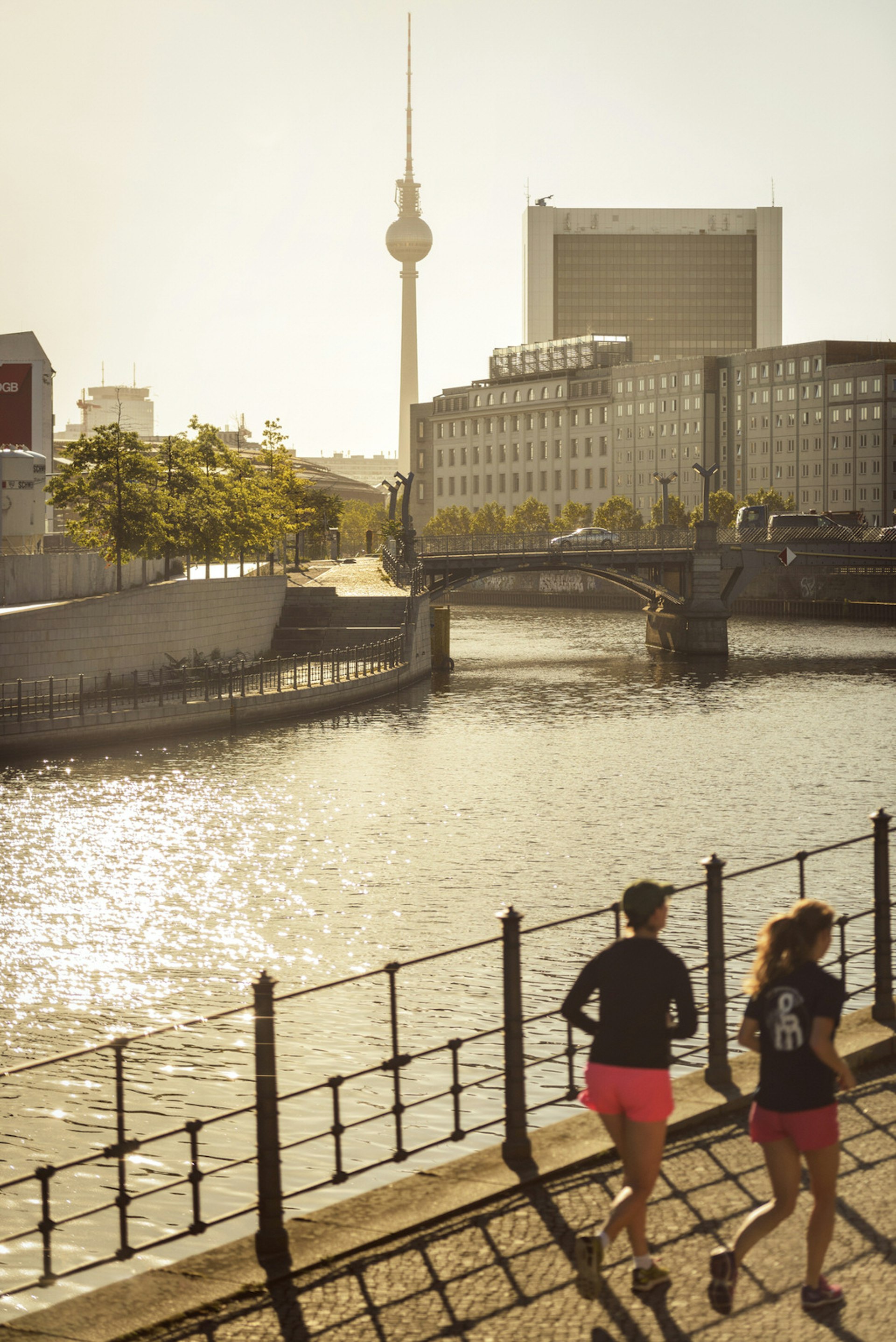 Two people are running at sunset along the paved bank of the hemmed in Spree River in Berlin, with the ever-present TV Tower looming in the distance
