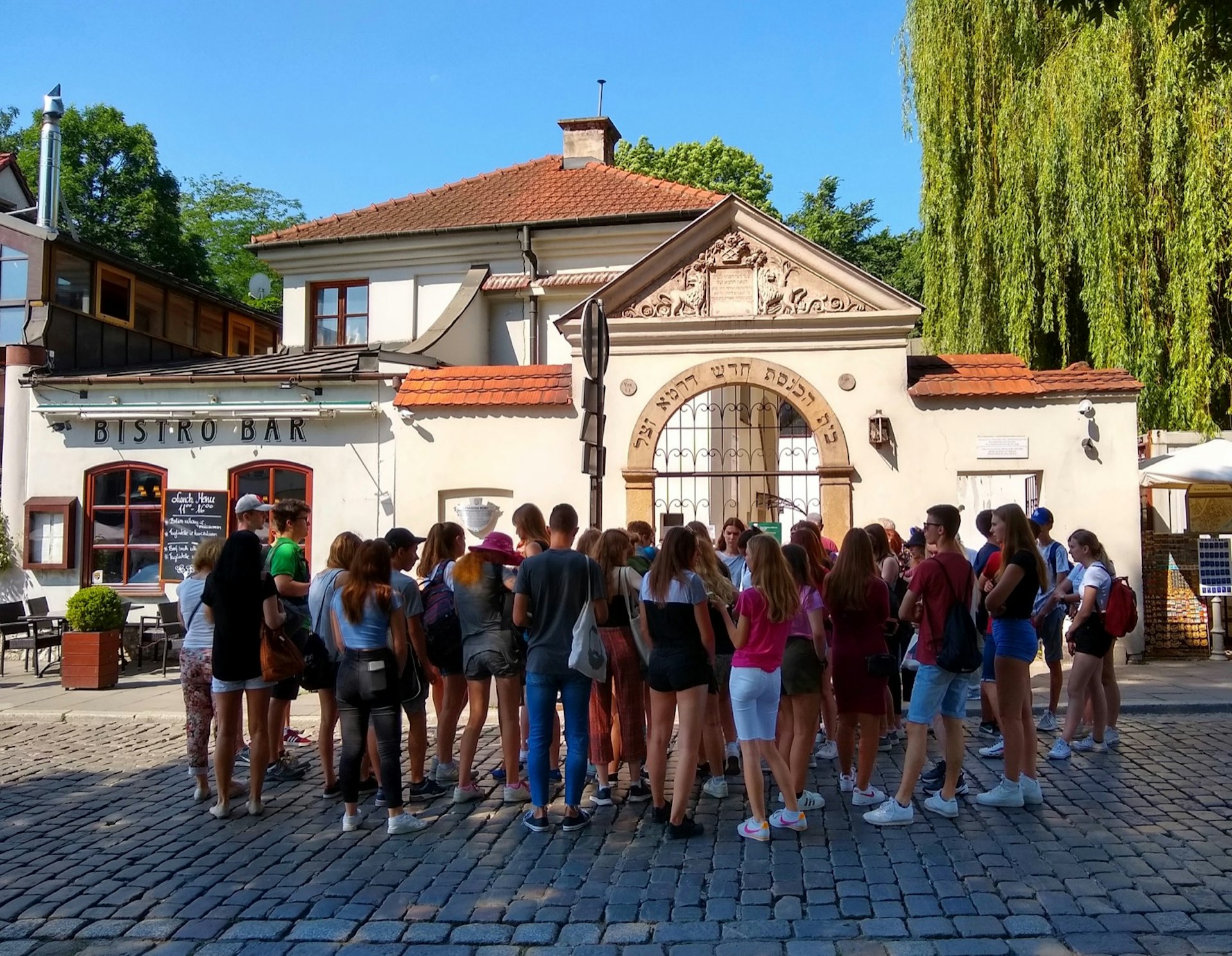 A large group of teenagers stands on a cobblestone street in front of a tan arch with Hebrew writing on it; Krakow Jewish culture revival