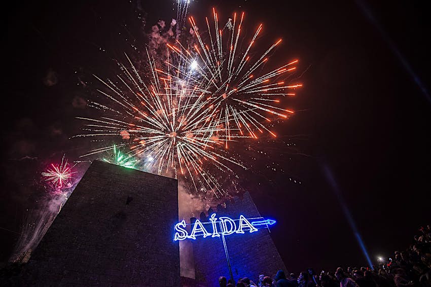 Fireworks lighting up the sky at the Sexta13 festival, above a neon "Saida" sign.