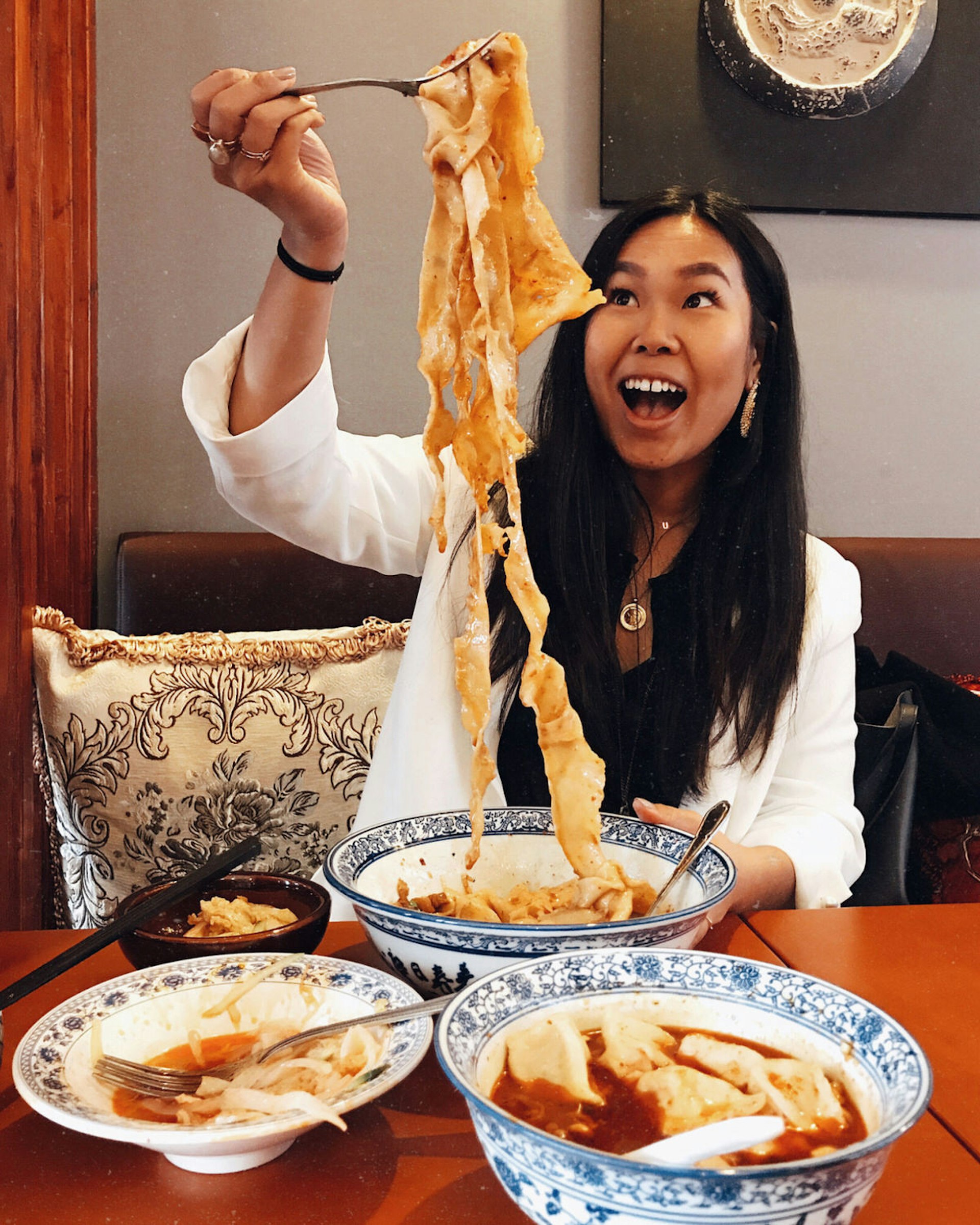 Shu Shi Lin raising pork biang biang noodles into the air from a bowl using a fork. Shu is looking up at the noodles in delight, there are two other saucy dishes on the table in white bowls with a blue pattern. 