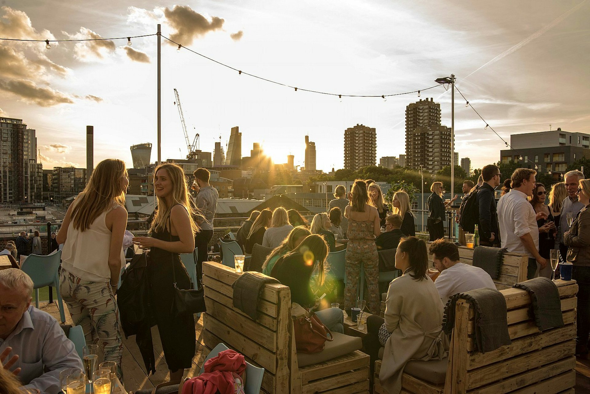 Drinkers converge at sunset on the Skylight rooftop; some are standing, while others sit on seats repurposed from wooden pallets, covered in grey cushions; the City of London and residential tower blocks are visible beyond.