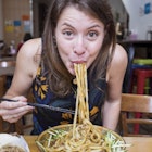 Sofia Levin biting into a mouthful of saucy noodles, looking into the camera with chopsticks in her hand. There are two other side dishes on the table and the restaurant is blurred in the background.