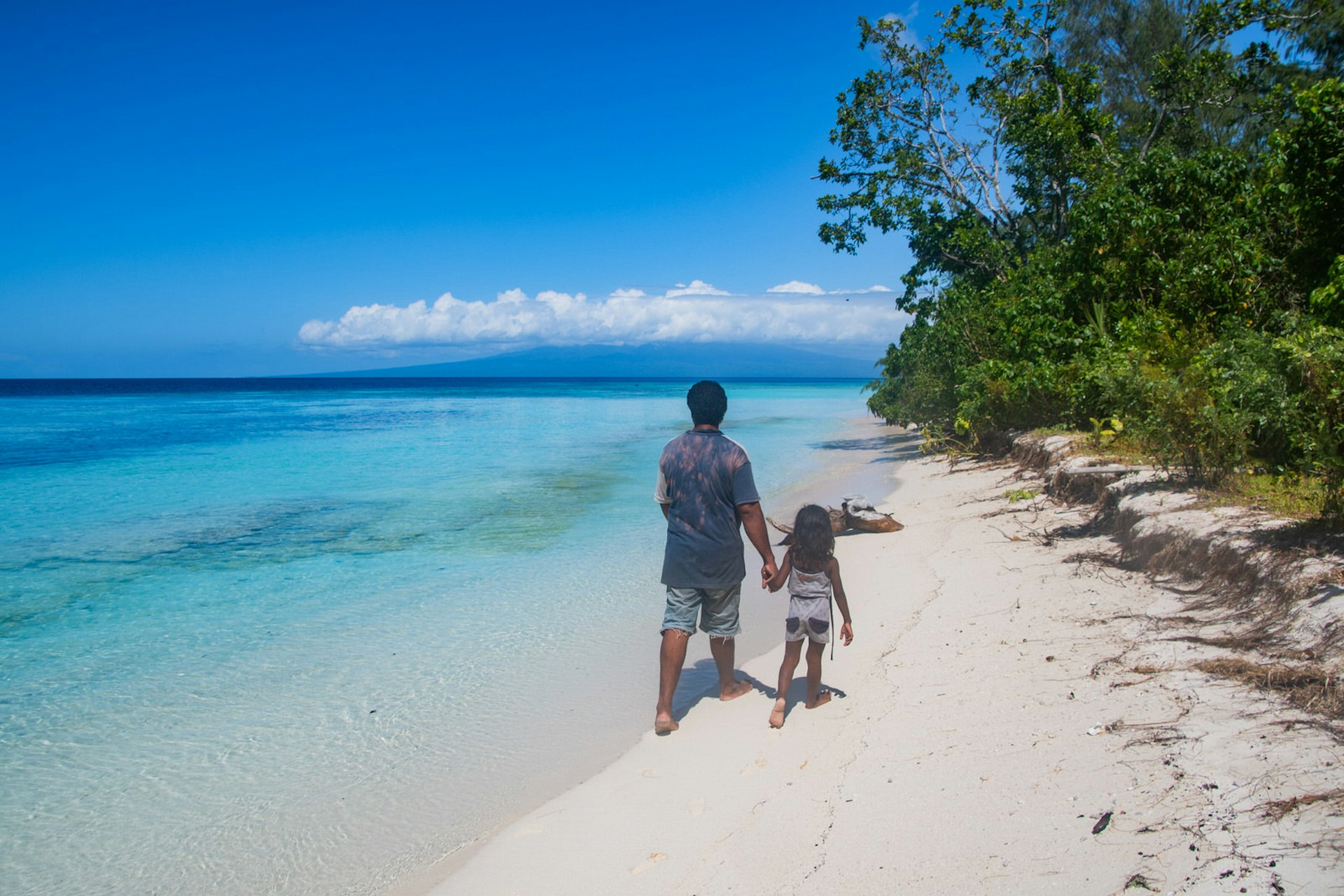 A man and his daughter walk along a pristine white sand beach holding hands. They are both wearing loose shorts and tops. There is one white cumulus cloud in an otherwise clear blue sky, clear sparkling water to their left and tropical greenery to their right. 