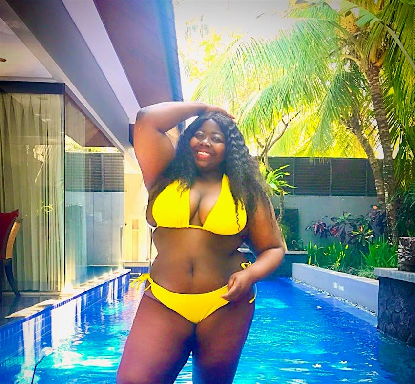 A black plus size woman poses in front of a private pool in a yellow bikini. The sun is streaming through the palm trees, she's smiling and looks really relaxed and happy. A black girl travelling in Bali tip: plus-size woman attract a lot of attention. 