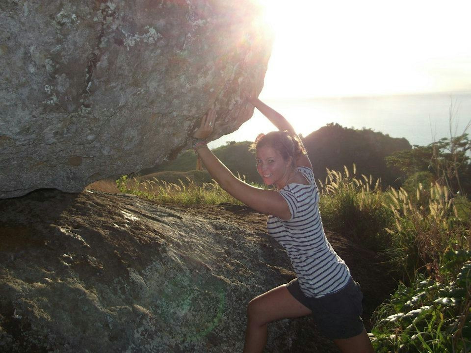 A smiling woman in a striped t-shirt and black shorts pushes against a huge boulder as the sun spills into the top of the frame.