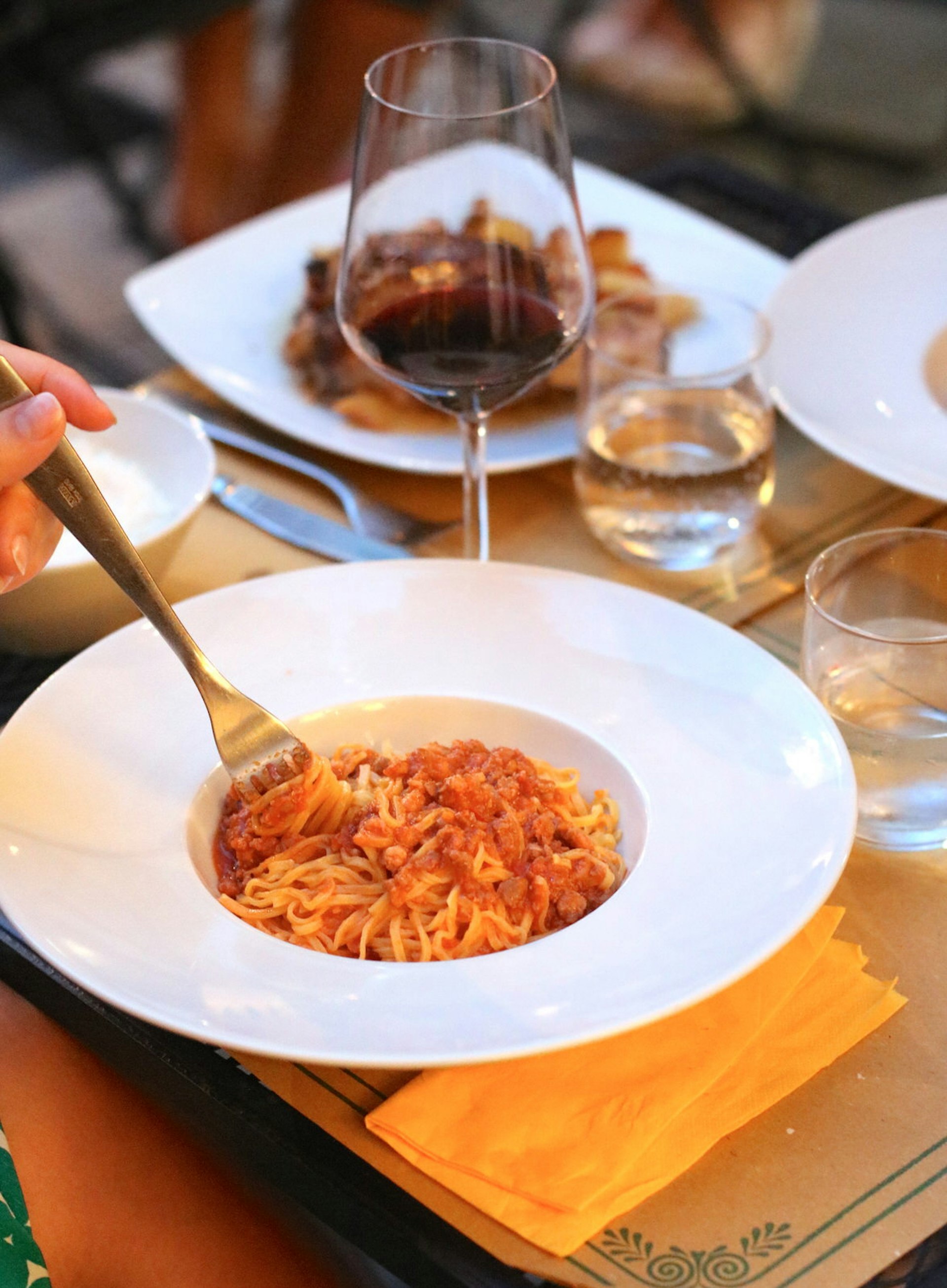 A close up shot of a bowl of Tajarin al sugo di carne - a meat and pasta dish. There are other dishes and a glass of red wine blurred in the background. 