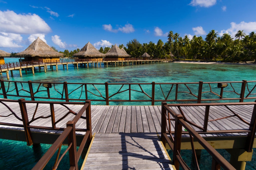 A view of several overwater bungalows from decking with clear turquoise water between the photographer and the bungalows.