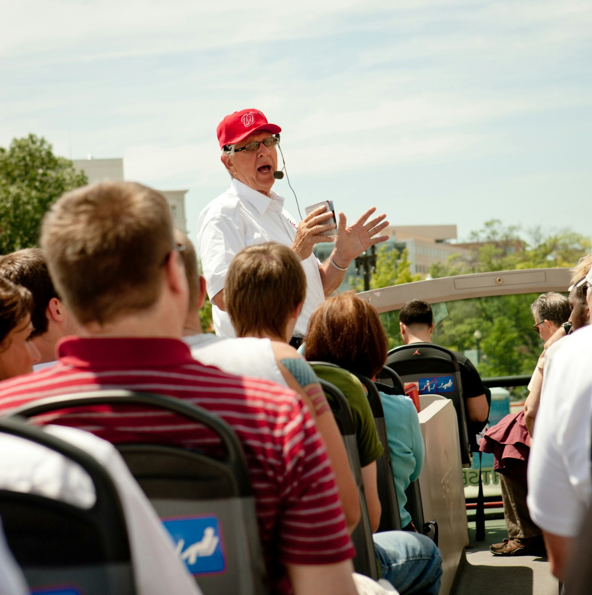 Tour guide talking about Washington to tourists on the top level of an open-air bus. The guide is wearing a red baseball cap and white shirt. The passengers backs are to the camera and it is a sunny day. 