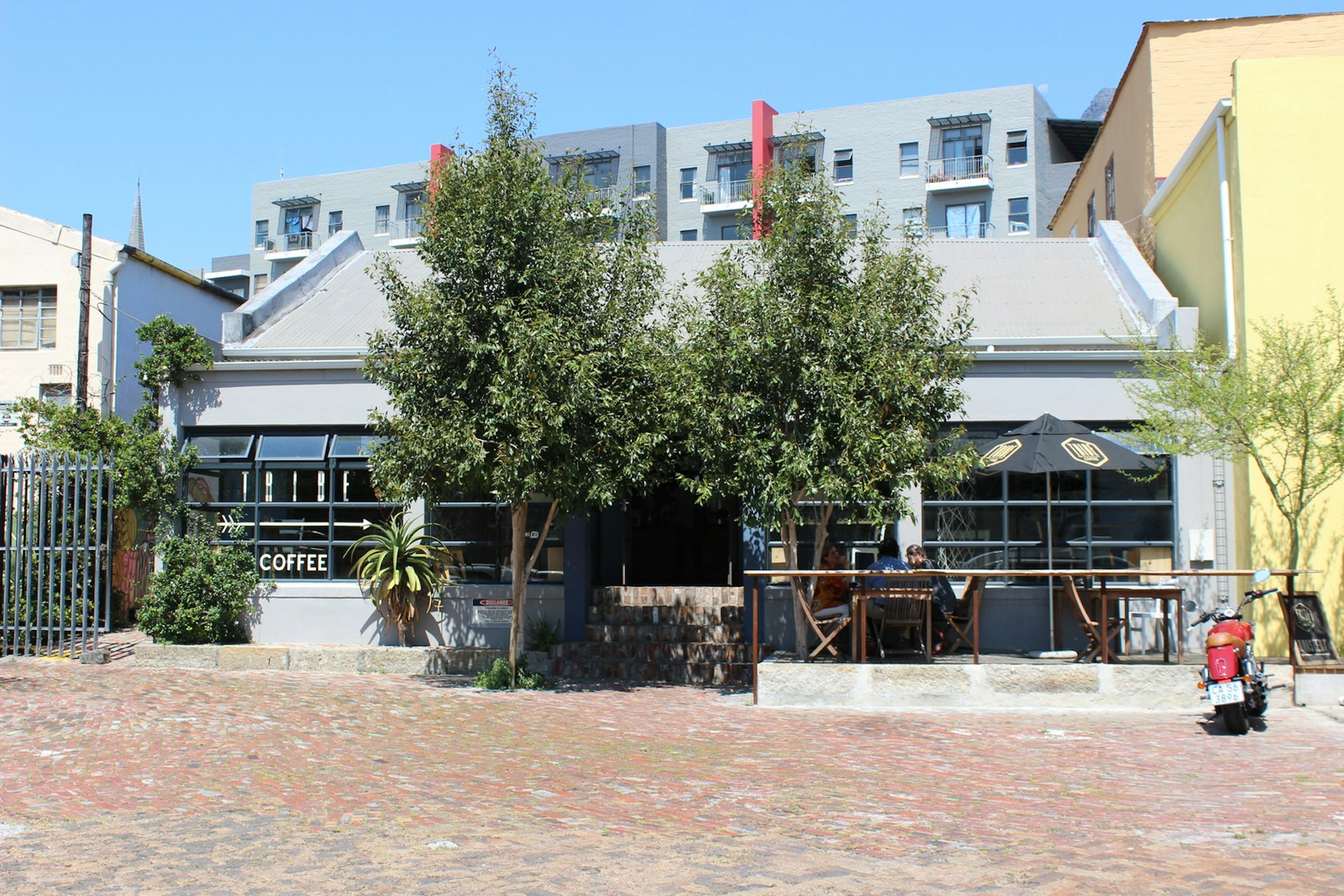 A grey, single storey building sits behind some trees on a wide cobbled street; there is an umbrella and some tables and chairs out front for patrons