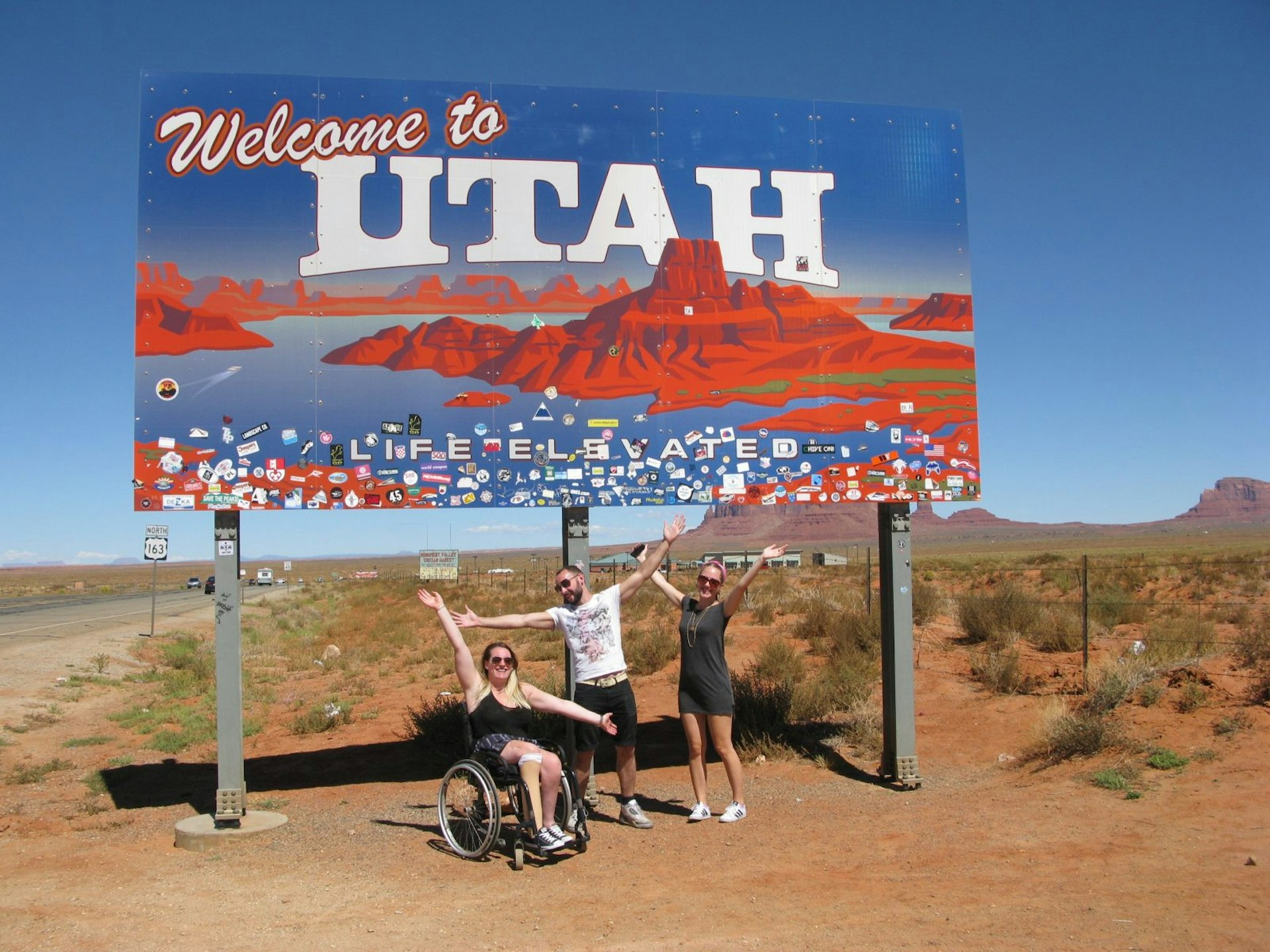 A large 'Welcome to Utah' sign is on the side of a dusty road. Three people, two women and one man, are underneath the sign with their arms outstretched in celebration. The woman on the left is in a wheelchair and the two others are standing next to her.
