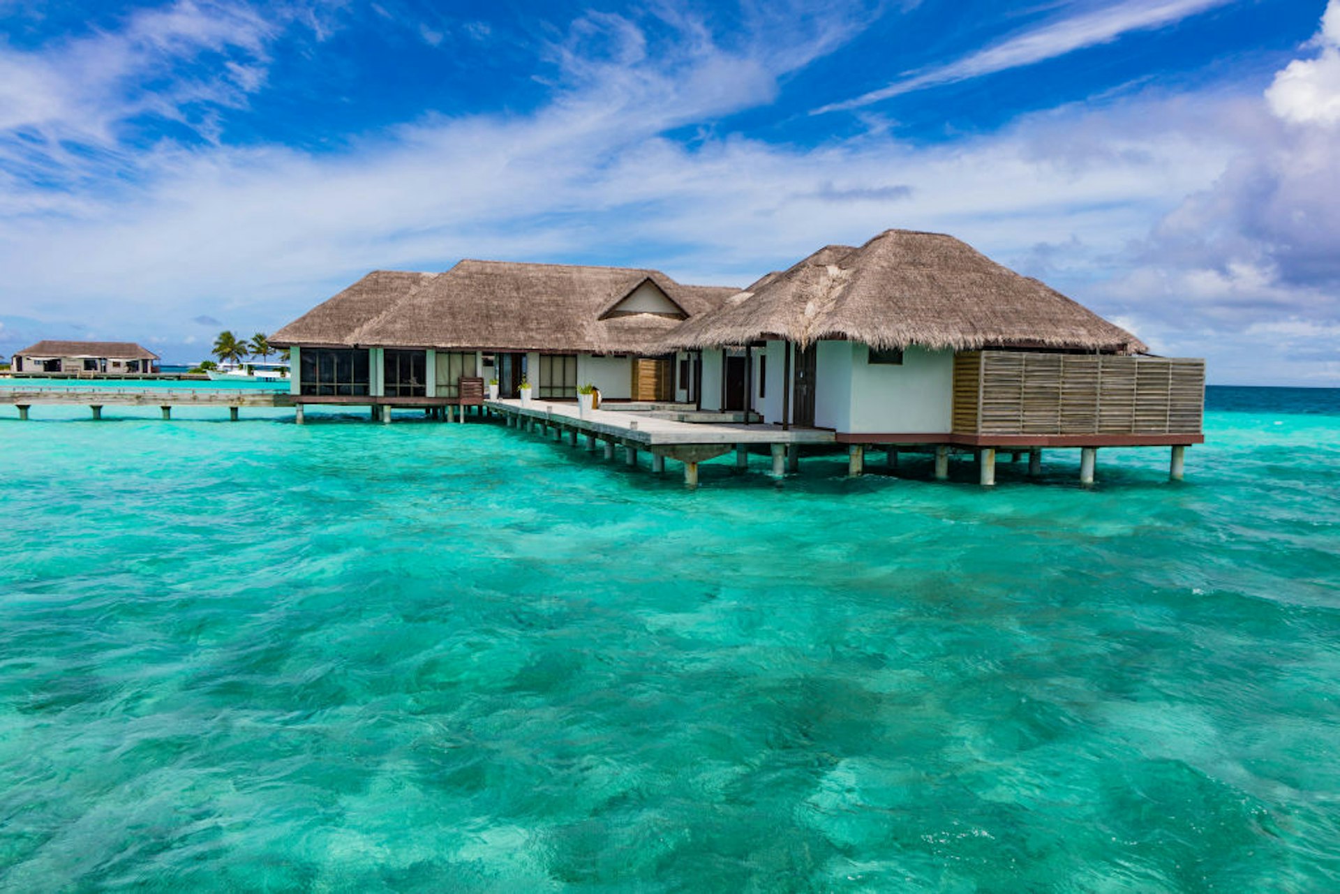Overwater bungalows in Velassaru Resort in the Maldives. Brown-thatch and white-walled cottages sit atop clear turquoise water.