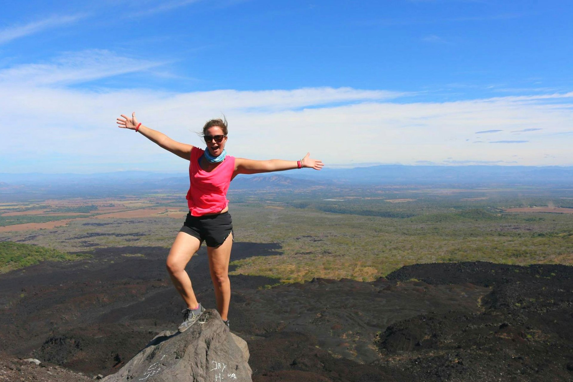 A woman stands on top of a volcano peak in trainers, black shorts and a pink sleeveless t-shirt with her arms outstretched. The Nicaraguan landscape spreads out far into the distance behind her and the blue sky is scattered with clouds.
