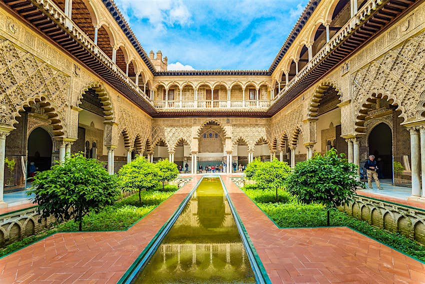 A courtyard inside Seville's Real Alcázar, surrounded by ornate gilded arches, plaster work and tiling with a sunken garden and long thin pool in the centre.