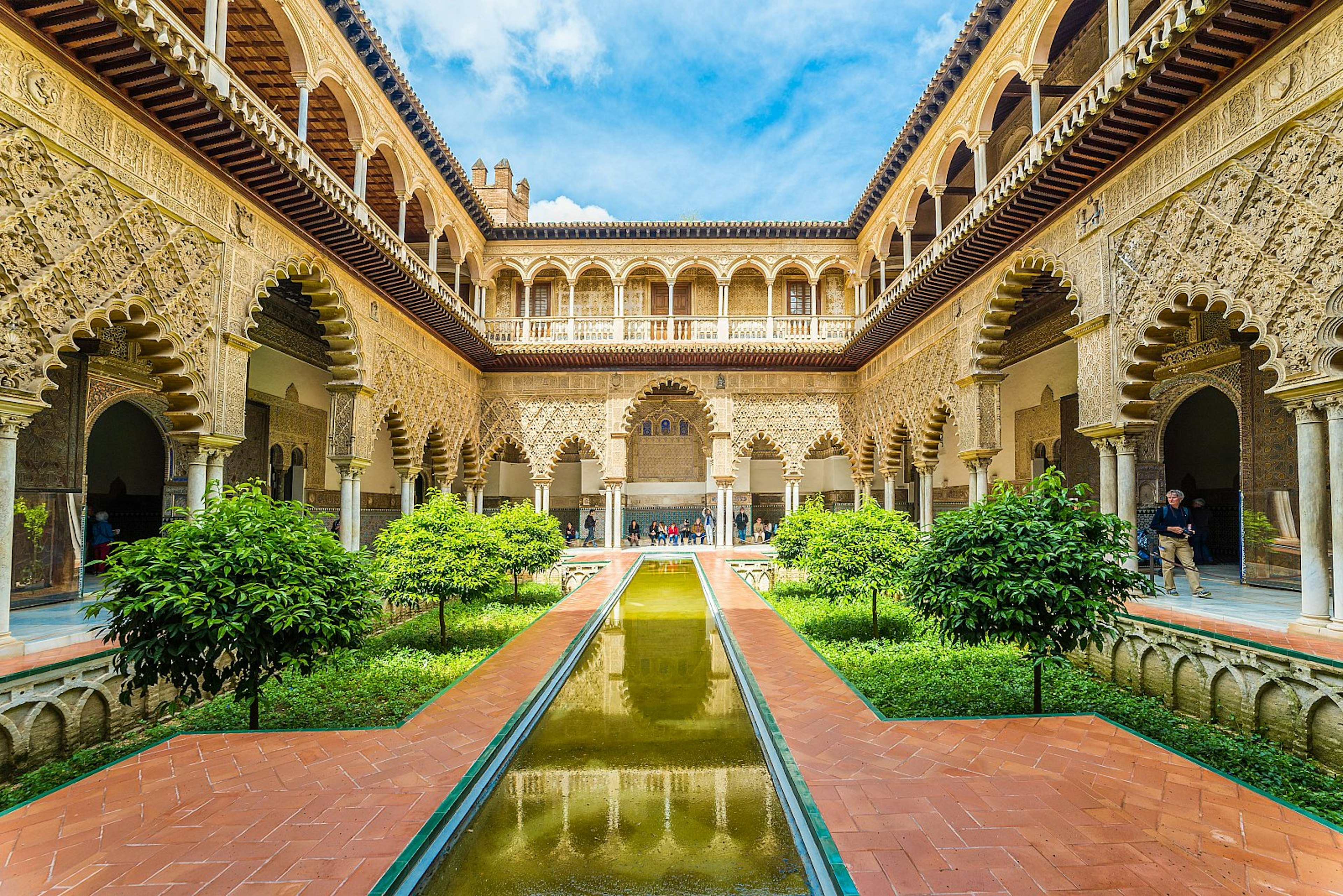 A courtyard inside Seville's Real Alcázar, surrounded by ornate gilded arches, plasterwork and tiling with a sunken garden and long thin pool in the centre.