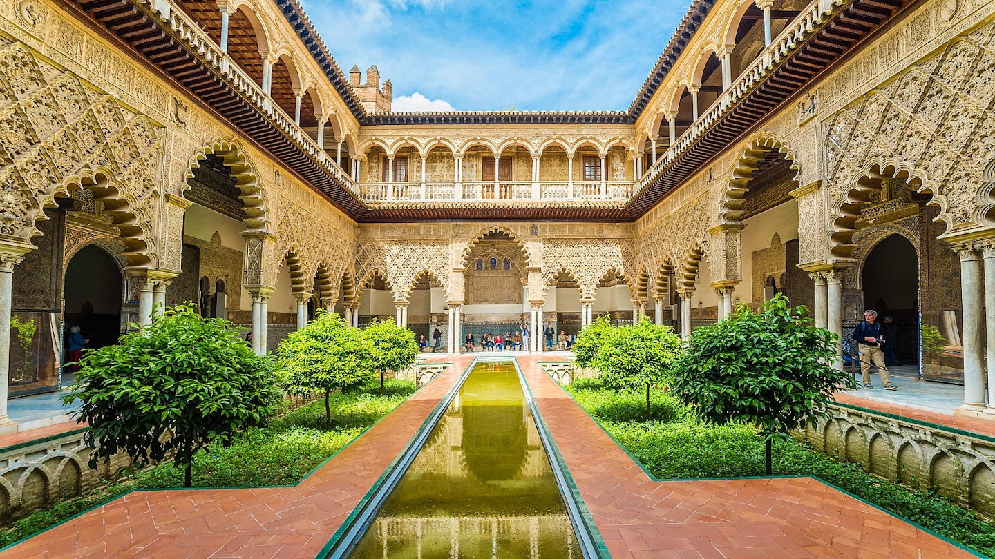 A courtyard inside Seville's Real Alcázar, surrounded by ornate gilded arches, plasterwork and tiling with a sunken garden and long thin pool in the centre.
