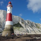 A group of people walking around the red and white lighthouse at eachy Head at low tide, with huge white cliffs rising behind it.