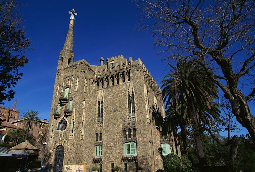 Features - bellesguard-barcelona-300b2e249e21 Bellesguard: it has a castle-like appearance with crenellated walls of stone and brick, narrow stained-glass windows, elaborate ironwork and a soaring turret mounted by a Gaudían cross. There are trees in its grounds.