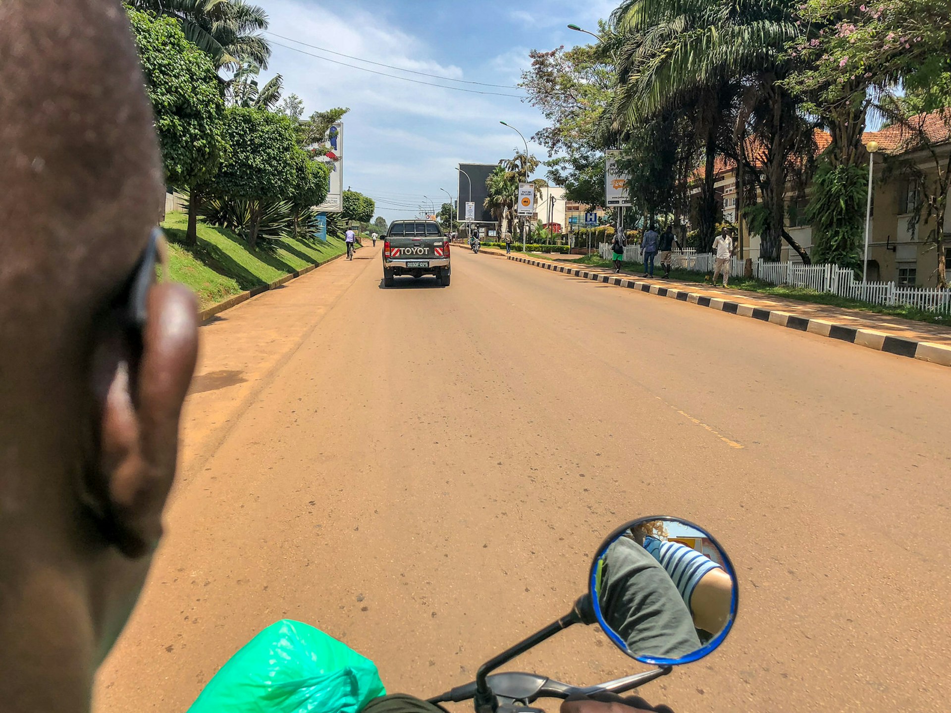 The right-hand side of the head of the motorcycle taxi (boda boda) driver is visible on the left side of the image; straight ahead is the sideview mirror and the paved road ahead