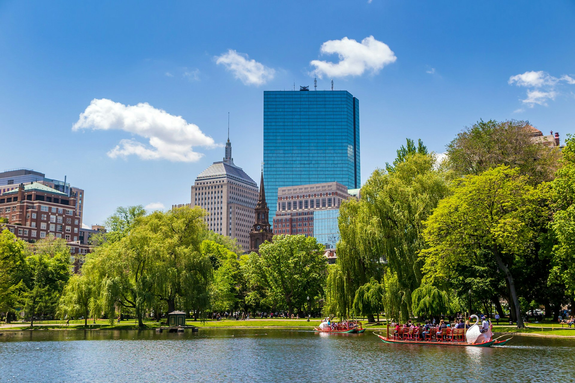 Visitors riding the swan boats at the Boston Public Garden on a sunny summer day; in the background, behind the tree-lined waterway are a couple of glass skyscrapers poking up into a blue sky dotted with a few whispy white clouds; baby travel
