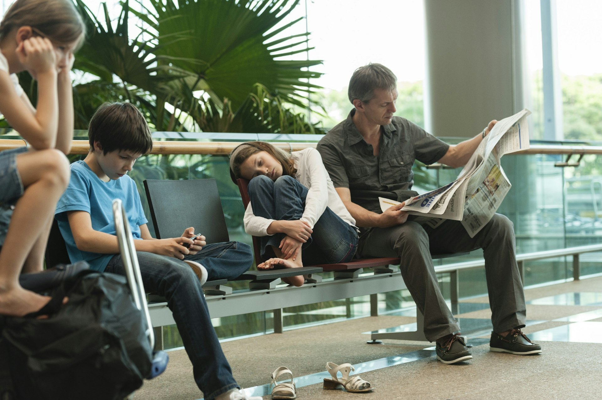 A family waits at the airport. A man reads the paper, one girl curls up on the seat, a boy plays with a phone while another girl sits on a suitcase with her head in her hands.