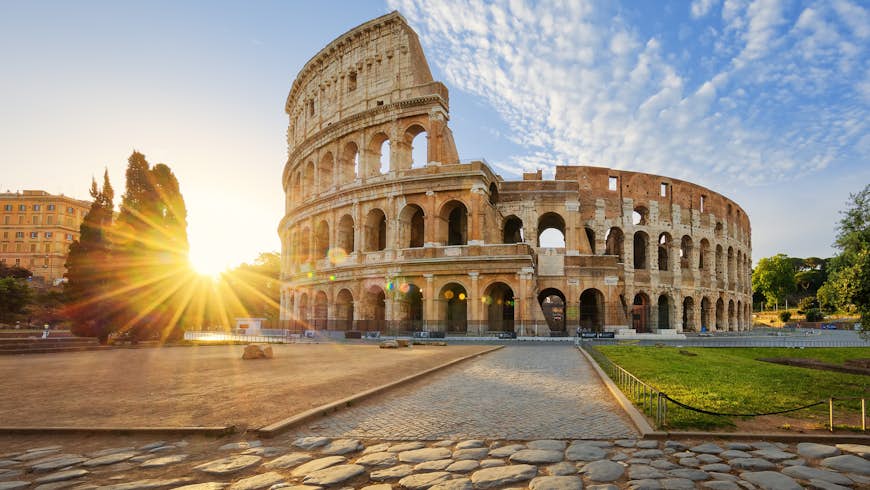 Looking over rough cobbles to a brilliant sun peaking over some distant trees, the Colosseum stands to the right in a golden light under a blue sky with cotton-like clouds above; an early start helps ensure a perfect weekend in Rome