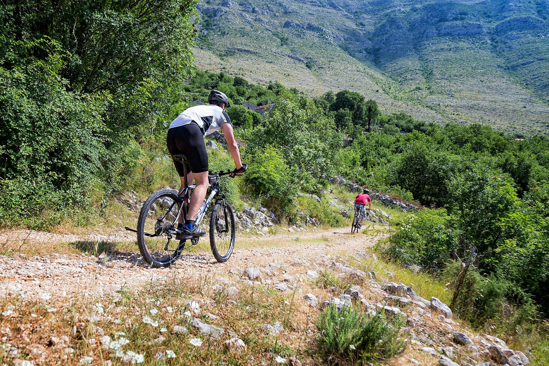 Two mountain bikers ride on a back-country road in Bosnia & Hercegovina; the track is surrounded by greenery and there is a rocky mountainside beyond.