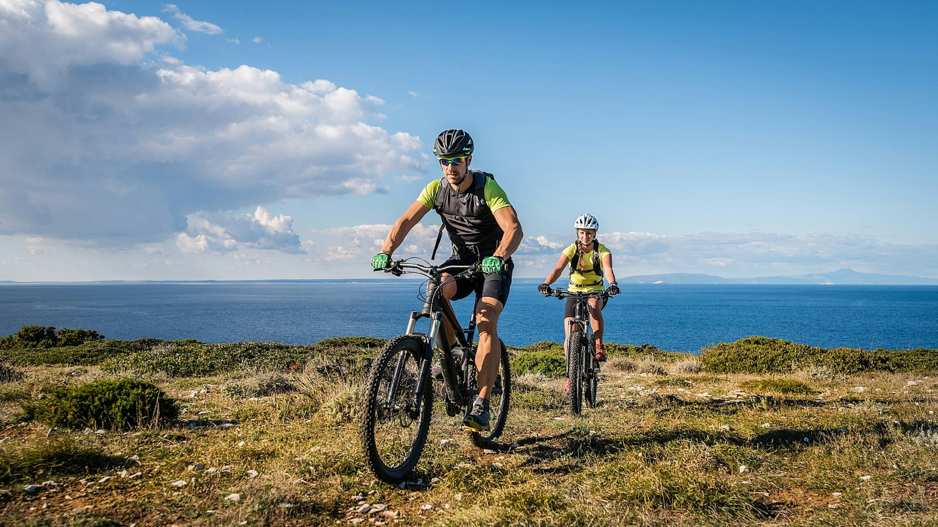 A few EuroVelo routes take in Croatia: here we see a young couple on mountain bikes cycling along a grassy headland, with the blue sea beyond.