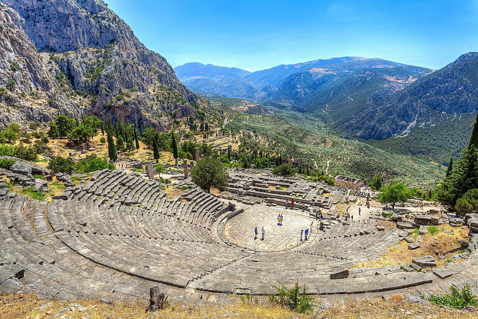 The well preserved remains of an ancient stone theatre on a mountainside; there is a lovely view down into a green valley below, surrounded by further mountain peaks.