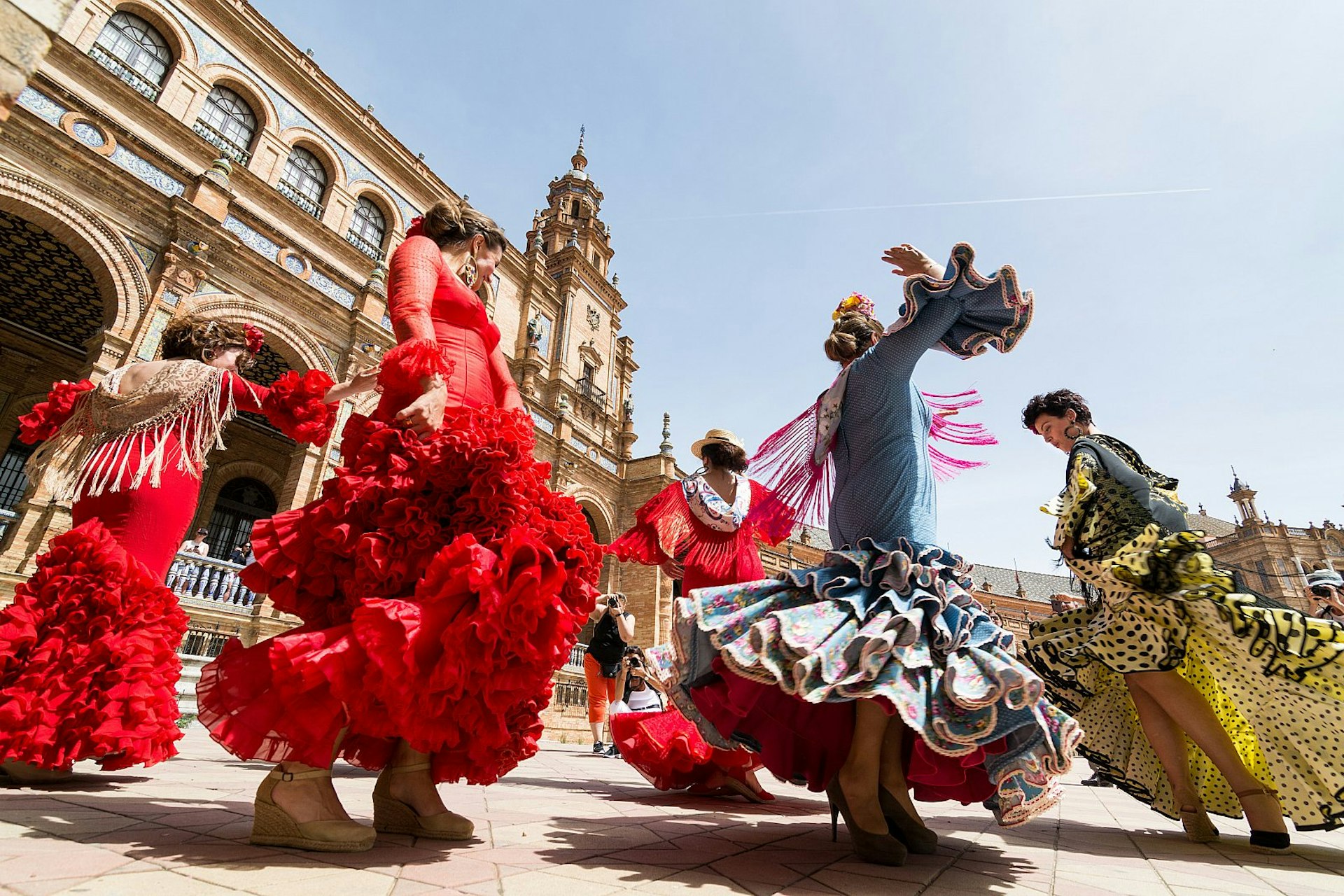 Kvinnor i färgglada klänningar dansar flamenco framför den storslagna arkitekturen på Plaza de España.