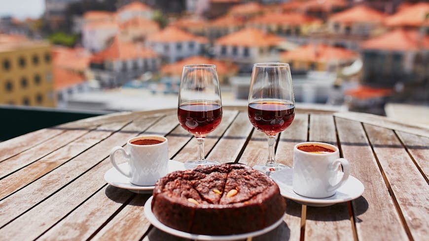 Two glasses of Madeira wine, two cups of espresso coffee and a traditional Portuguese honey and nut cake bolo de mel sit on a wooden tabletop with a view beyond of the blurred terracotta orange roofs of Funchal.