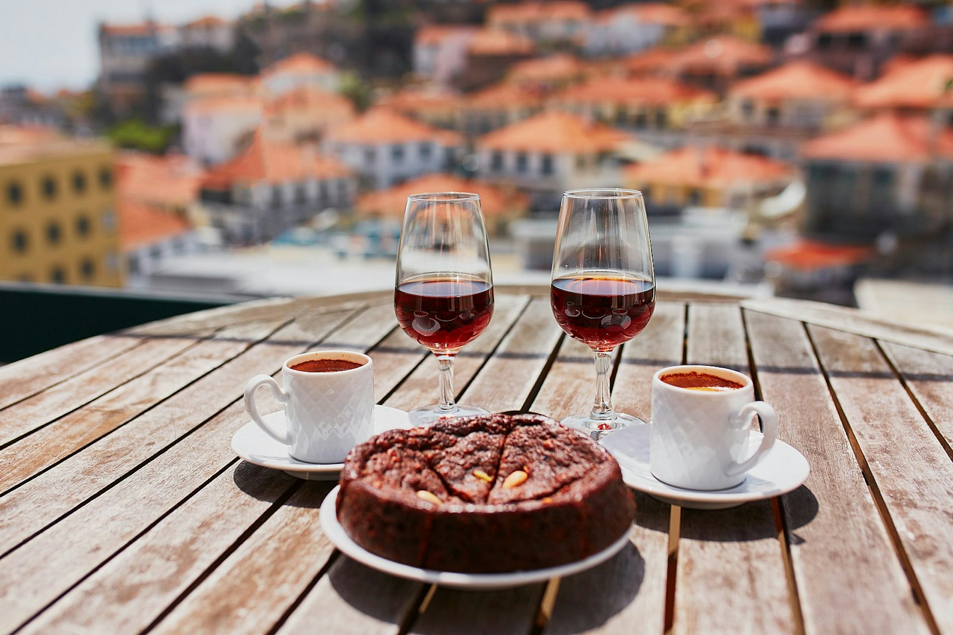 Two glasses of Madeira wine, two cups of espresso coffee and a traditional Portuguese honey and nut cake bolo de mel sit on a wooden tabletop with a view beyond of the blurred terracotta orange roofs of Funchal.