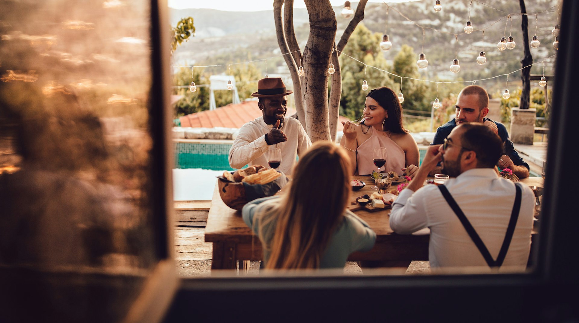 A group of five ethnically diverse people sit at a table in the sunshine eating and chatting.