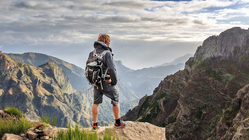 A hiker stands on a rocky ledge looking out over the rugged mountain scenery of the Pico do Arieiro in Madeira. They wear a waterproof jacket, back pack and shorts in shades of grey.