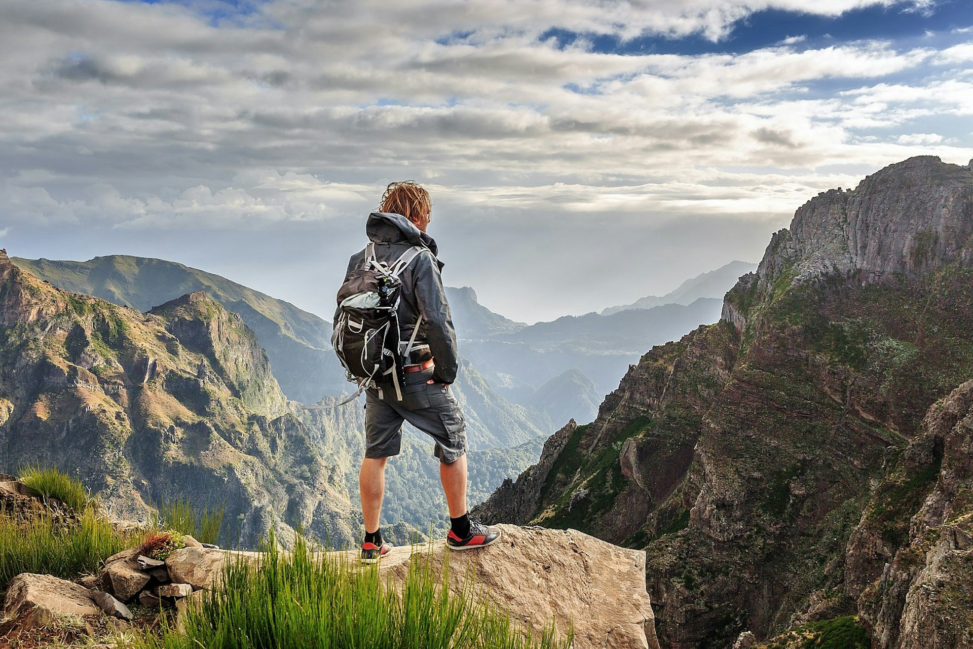 A hiker stands on a rocky ledge looking out over the rugged mountain scenery of the Pico do Arieiro in Madeira. They wear a waterproof jacket, back pack and shorts in shades of grey.