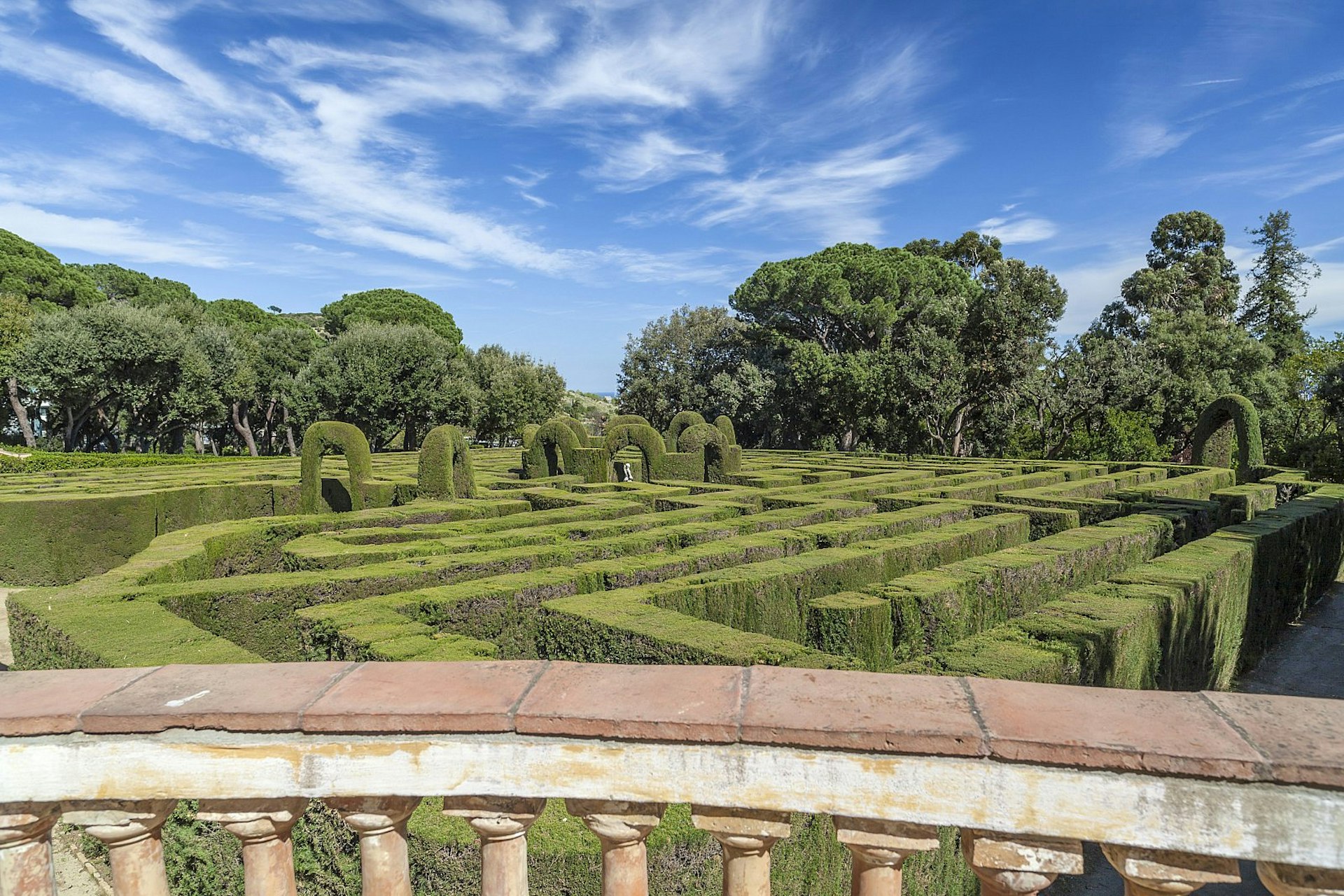 Looking over a pillared wall to a cypress hedge labyrinth below, with trees beyond.