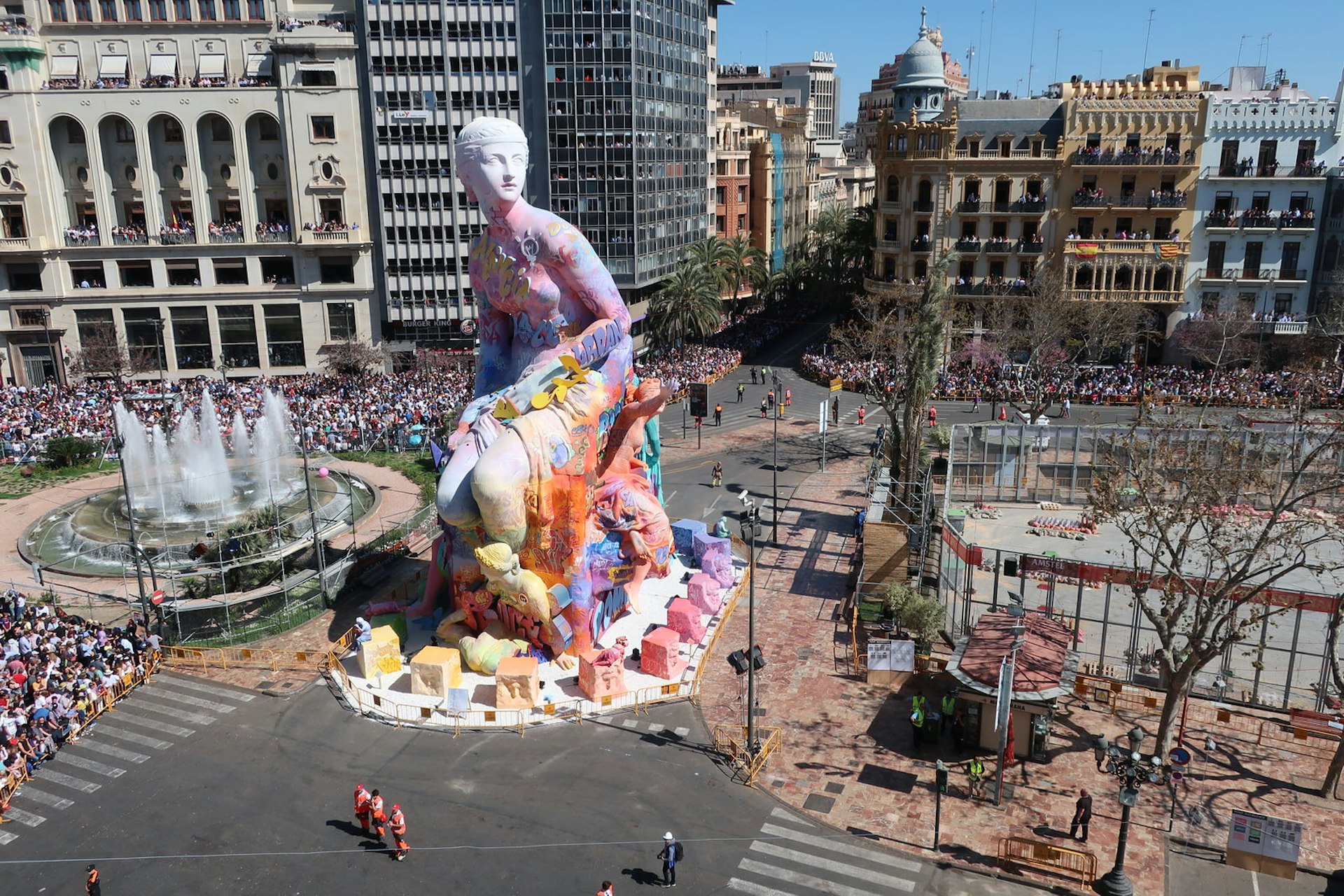 A massive, neoclassical-style sculpture of a woman, heavily covered in graffiti, stands in the middle of a large plaza, where crowds have gathered.