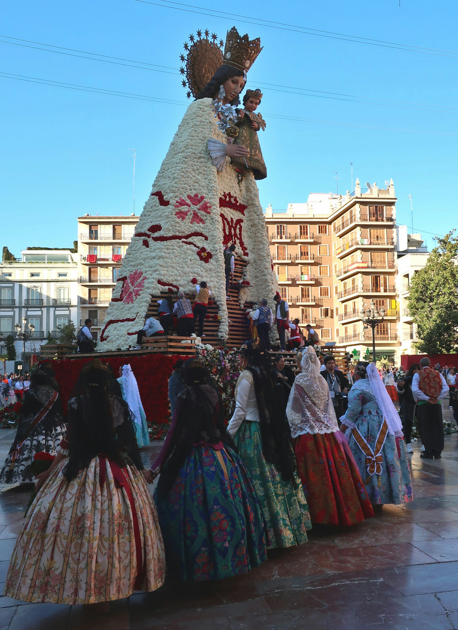 Five women in traditional dress look up at a wooden statue of the Virgin Mary, which is being decorated with bunches of flowers