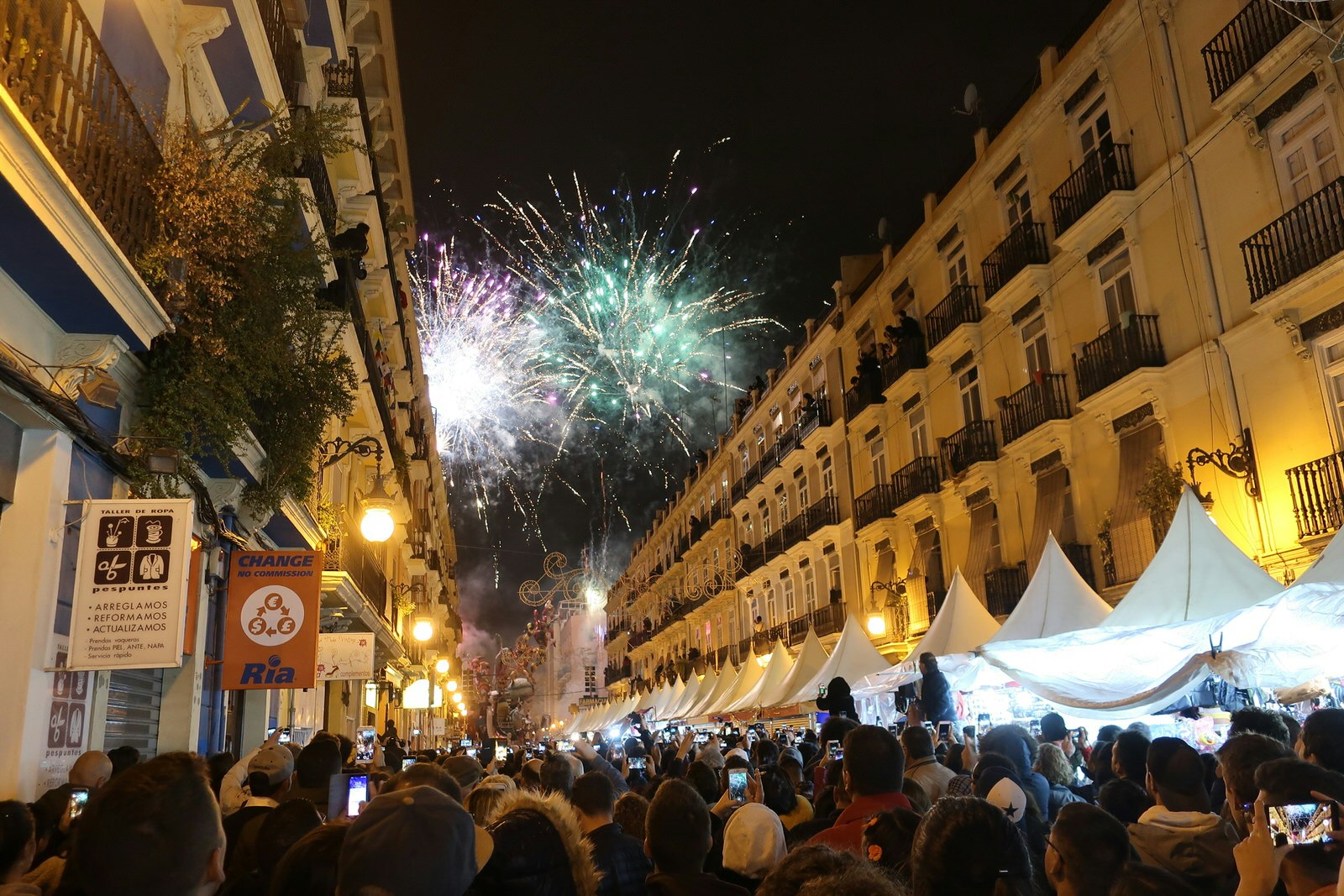 A crowd of people fill a street beneath tall buildings, while fireworks explode overhead.