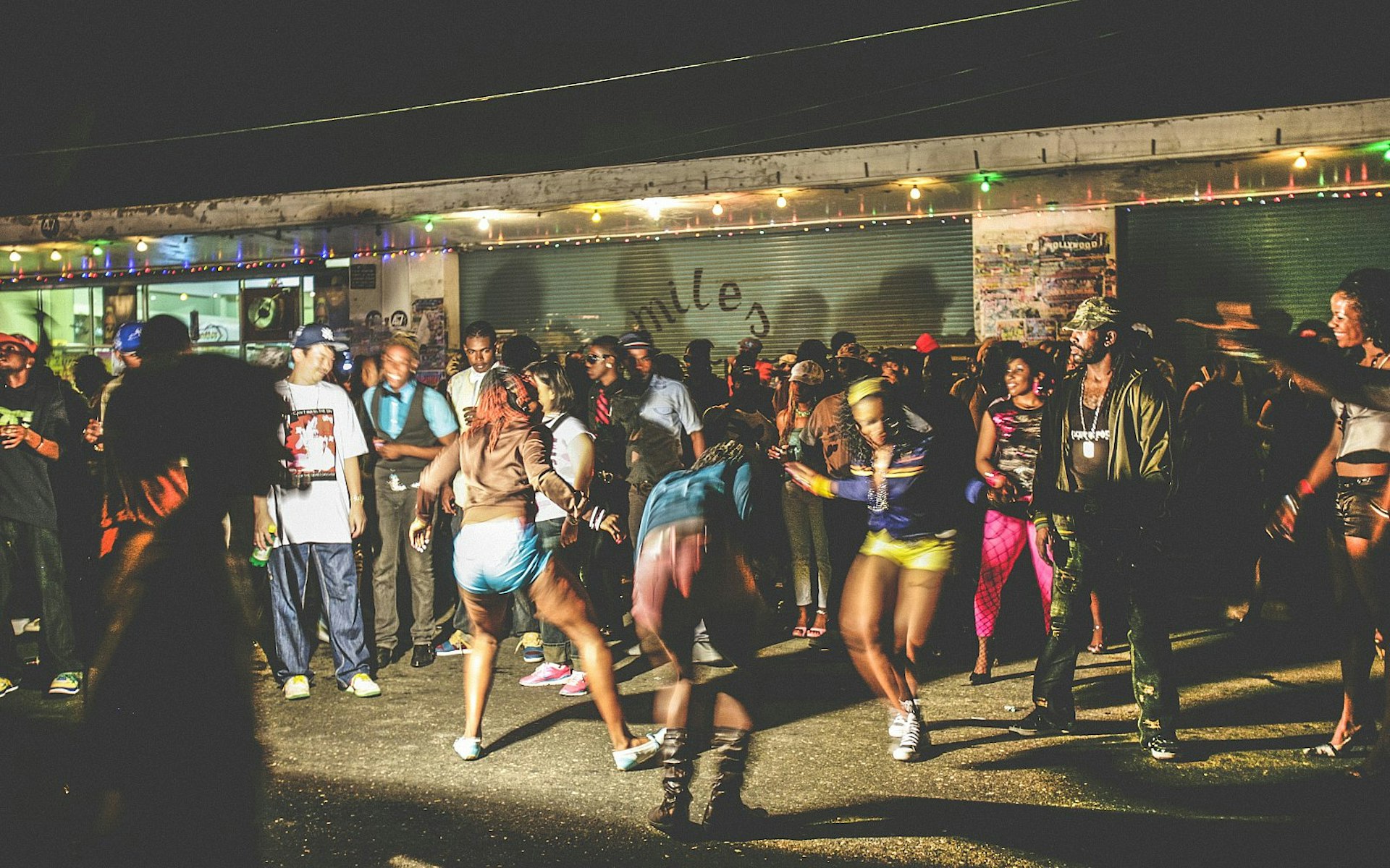 A group of people dancing in the street in Kingston, Jamaica, at night-time, next to a shuttered shop.