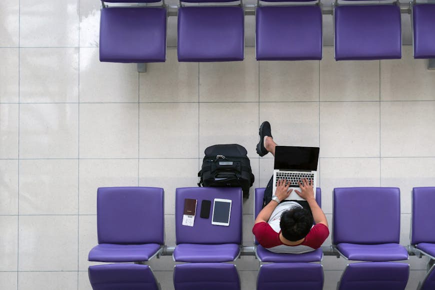 En flygbild av en manlig resenär som bär shorts och t-shirt arbetar på sin bärbara dator i en avgångslounge på flygplatsen.  Hans surfplatta, telefon och pass ligger på en lila sits bredvid honom och hans väska ligger på marken.